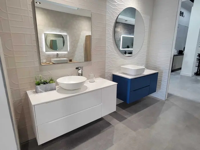 Bathroom vanity showroom display featuring two modern sink configurations with white wavy tile walls, LED-lit round mirrors, white and blue vanity cabinets, and vessel sinks on quartz countertops