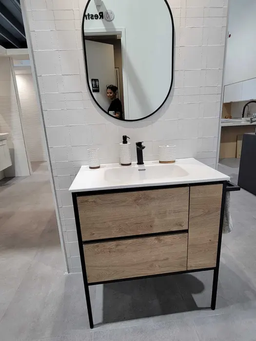 Modern bathroom vanity display with oak wood cabinetry, white countertop, black metal frame, oval mirror, and black matte faucet fixtures in a showroom setting.