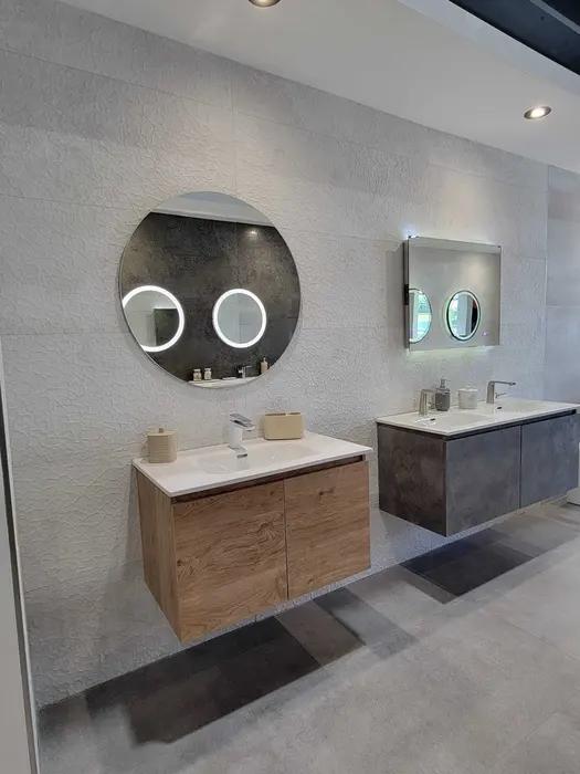 Bathroom vanity showroom display featuring two side-by-side sink stations with contrasting wood finishes, illuminated round mirrors, and modern fixtures against textured white tile walls