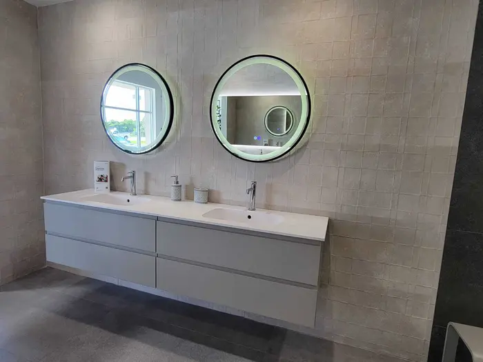 Completed master bathroom vanity with dual sinks, modern lighting, and tiled walls featuring two illuminated round mirrors and chrome faucets