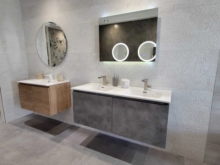 Modern bathroom vanity showroom display featuring two sink configurations: a natural wood cabinet on the left with round mirror and a dark grey cabinet on the right with dual LED-lit circular mirrors, white countertops, and textured white wall tiles.