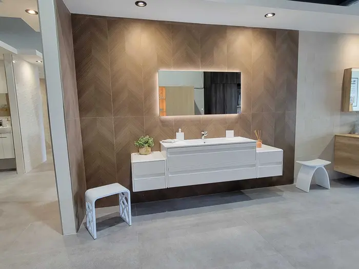 Modern bathroom vanity display with floating white cabinetry, herringbone tile accent wall, integrated mirror lighting, and decorative accessories in a showroom setting