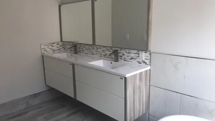 Completed bathroom vanity with dual sinks, white quartz countertop, glass tile backsplash, contemporary faucets, and large mirrors in a freshly renovated bathroom.