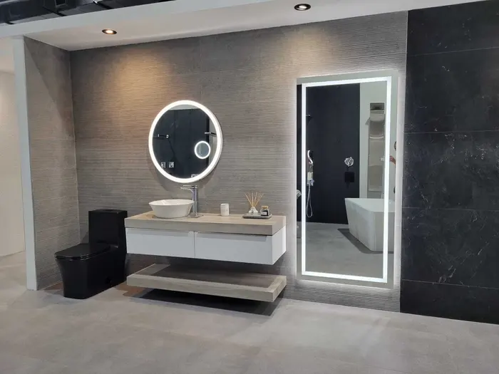 Modern bathroom with textured gray tile walls, floating white vanity with round LED mirror, black toilet, and partial view of walk-in shower with black tile