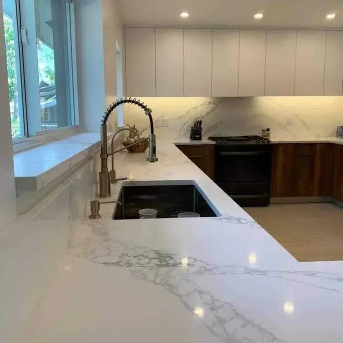 Completed modern kitchen with white marble countertops, black kitchen sink with chrome faucet, wood-grain cabinetry, white upper cabinets, marble backsplash, recessed lighting, and natural light from large windows