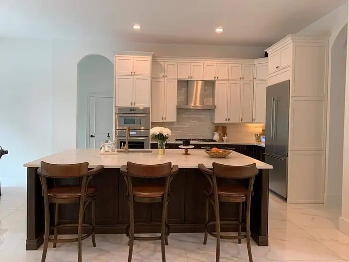 Modern kitchen with cream-painted cabinetry, stainless steel appliances, subway tile backsplash, and a dark wood island with beige countertops and three wooden bar stools