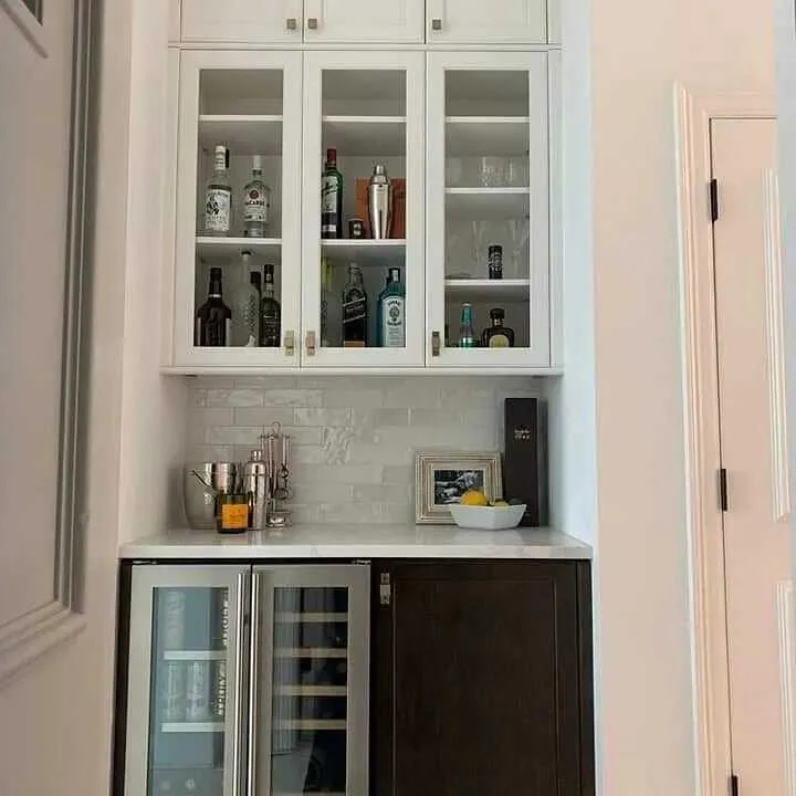 Completed modern kitchen bar with white glass-front upper cabinets displaying spirits, gray subway tile backsplash, white quartz countertop, and dual wine/beverage cooler below