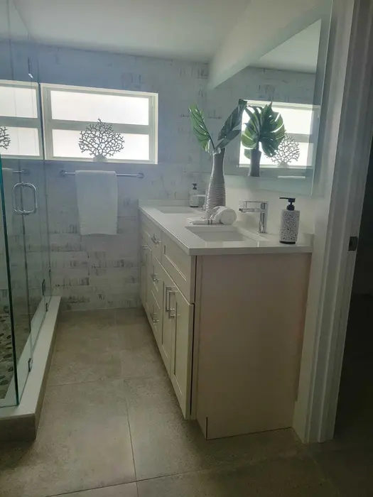 Modern completed bathroom with white tile shower surround, light wood vanity with white countertop, chrome fixtures, and decorative windows with natural light
