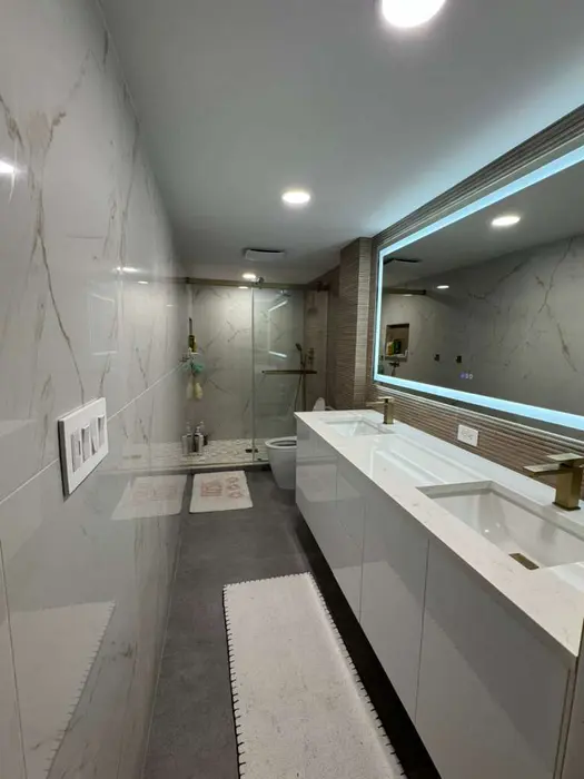 Completed modern bathroom with marble tile walls, large LED-backlit mirror, floating white vanity with dual sinks, walk-in shower with glass enclosure, and recessed lighting