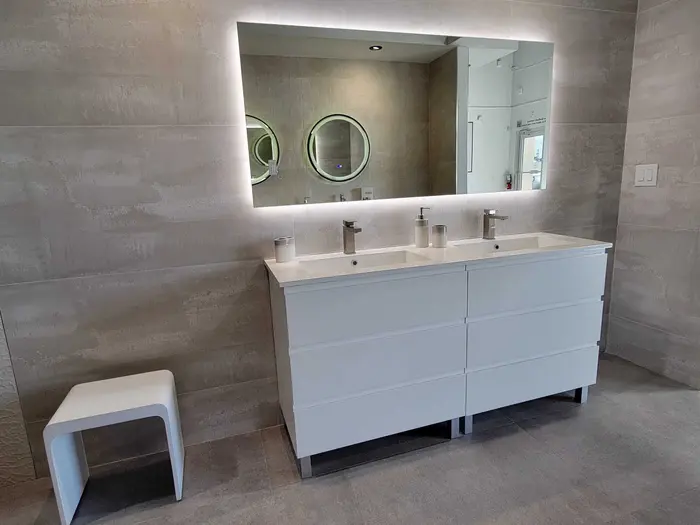 Baño moderno con tocador blanco doble, espejo retroiluminado, azulejos tipo cemento en las paredes y banqueta a juego