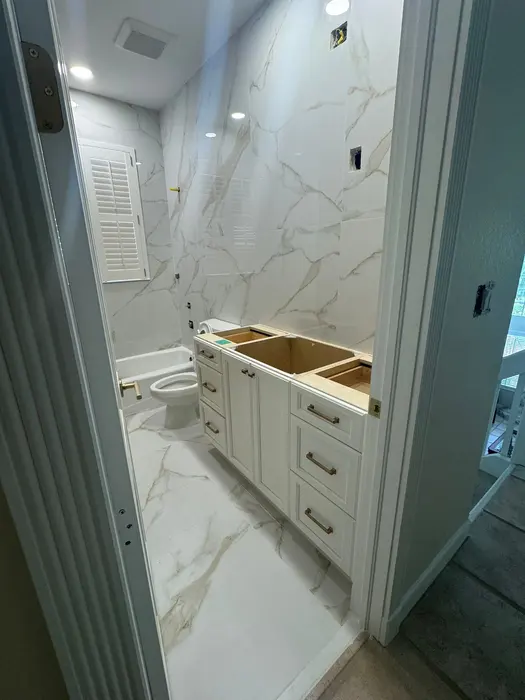 Un baño durante la renovación mostrando paredes y piso con azulejos de mármol, base de gabinetes blancos sin encimera, iluminación empotrada e instalación de inodoro en progreso.
