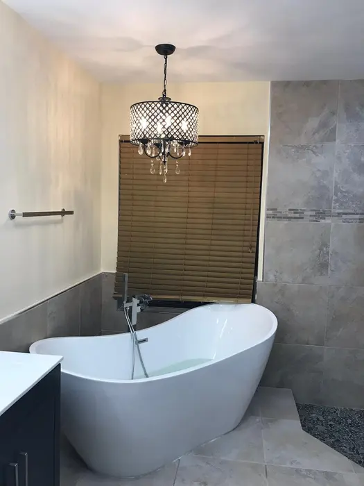Baño moderno completado con tina ovalada blanca independiente, paredes de azulejos grises, luminaria colgante bronce con detalles de cristal, y cortina de ventana de lamas de madera
