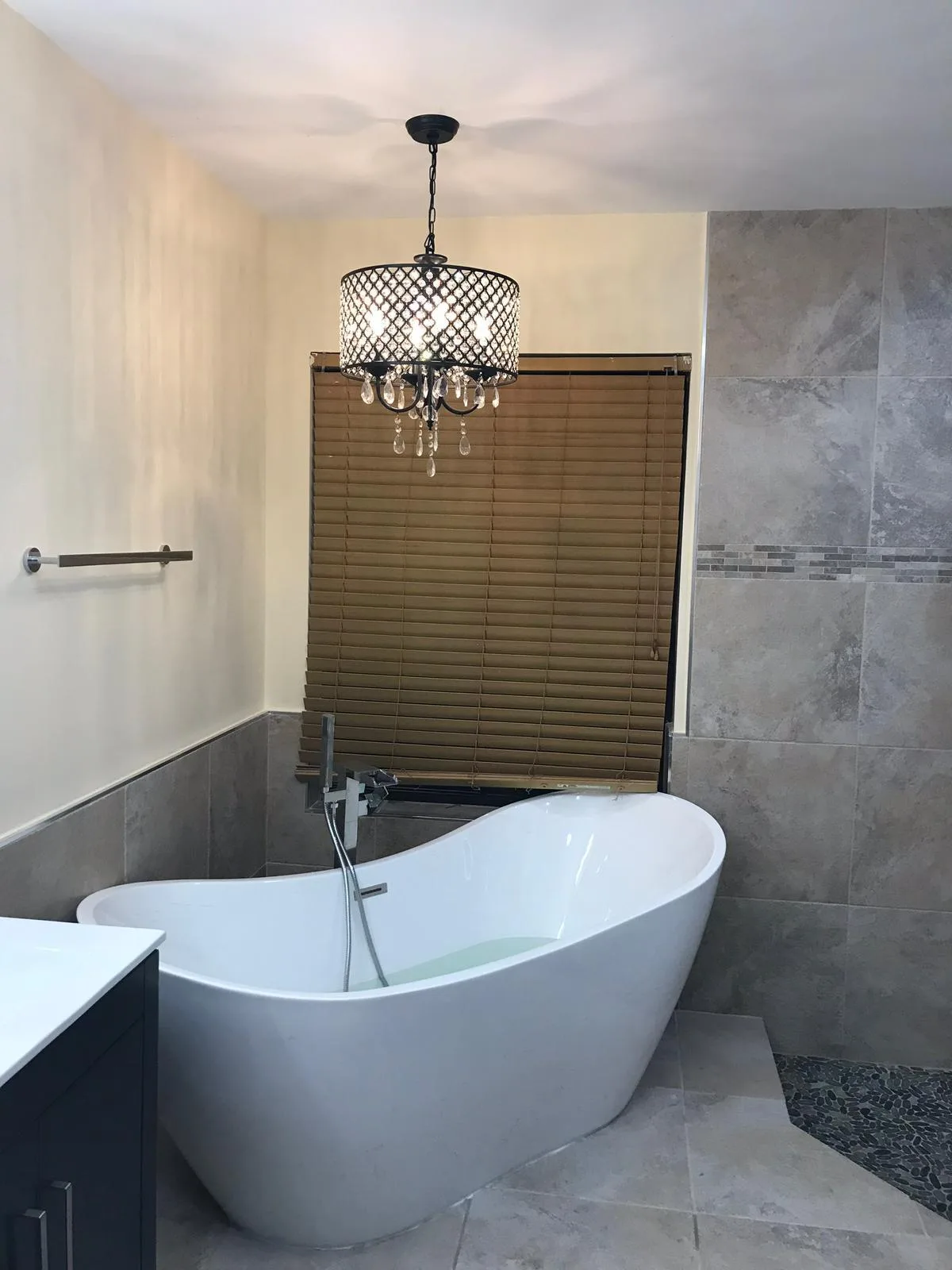 Baño moderno completado con tina ovalada blanca independiente, paredes de azulejos grises, luminaria colgante bronce con detalles de cristal, y cortina de ventana de lamas de madera