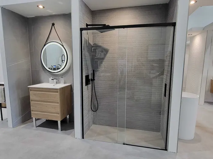 Baño moderno con tocador de madera clara, espejo redondo con marco negro, paredes de azulejos gris, y ducha abierta con cerramiento de vidrio