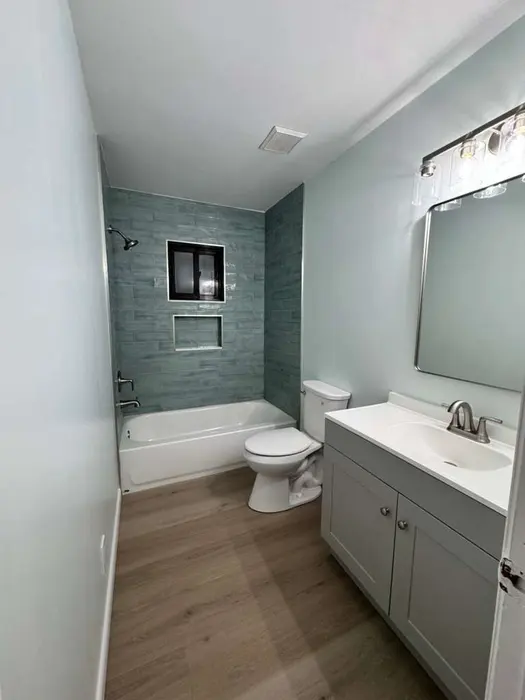 Baño moderno con mampara de ducha revestida en azulejos gris pizarra, vanidad blanca con lavamanos integrado y piso tipo madera