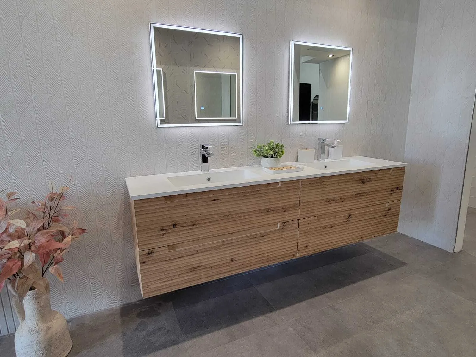 Vanidad de baño moderna completada con doble lavamanos, espejos iluminados y gabinetes de acabado grano de madera contra paredes de azulejo gris texturado.