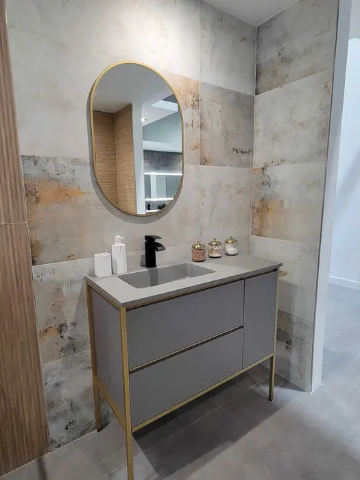 Mueble de baño moderno con gabinetes grises, herrajes de latón, lavamanos con encimera blanca y espejo ovalado con marco de latón sobre paredes con azulejos texturizados