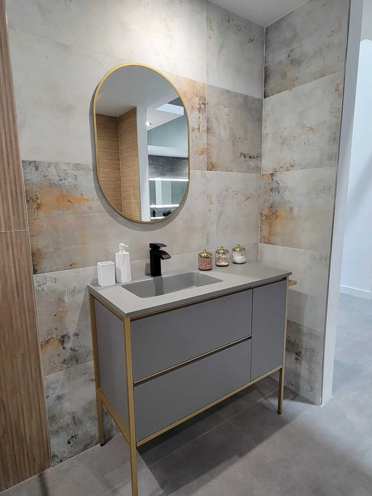 Mueble de baño moderno con gabinetes grises, herrajes de latón, lavamanos con encimera blanca y espejo ovalado con marco de latón sobre paredes con azulejos texturizados