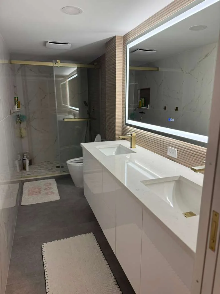 Baño moderno terminado con doble lavabo blanco, pared de tocador con acento de madera, espejo con luz LED, nicho de ducha con azulejos de mármol y accesorios de iluminación contemporáneos