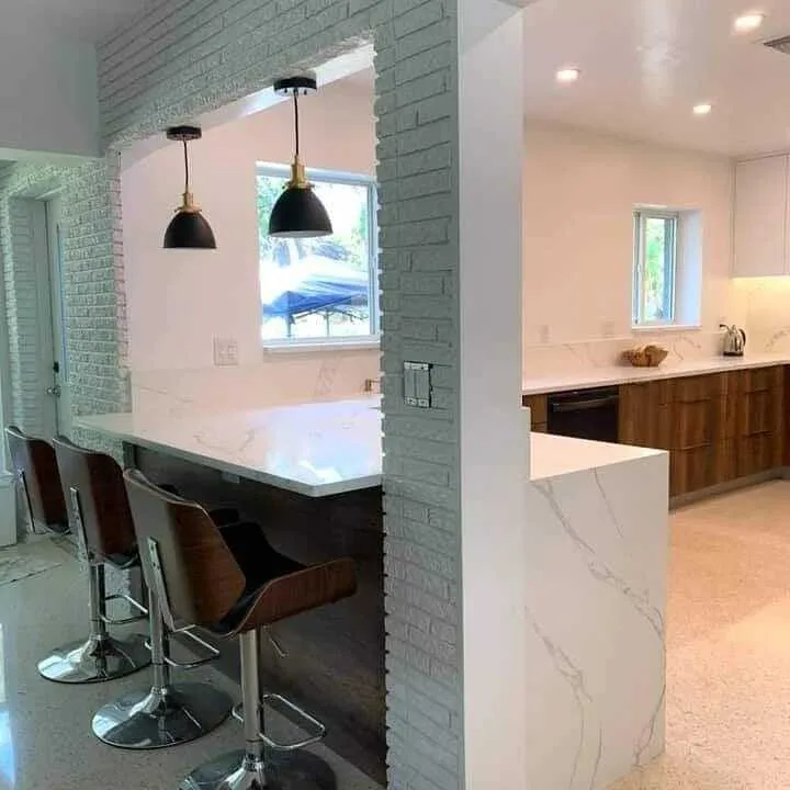 Cocina moderna completada con isla de cubierta mármol blanco, pared de ladrillo verde, iluminación colgante y gabinetes de madera
