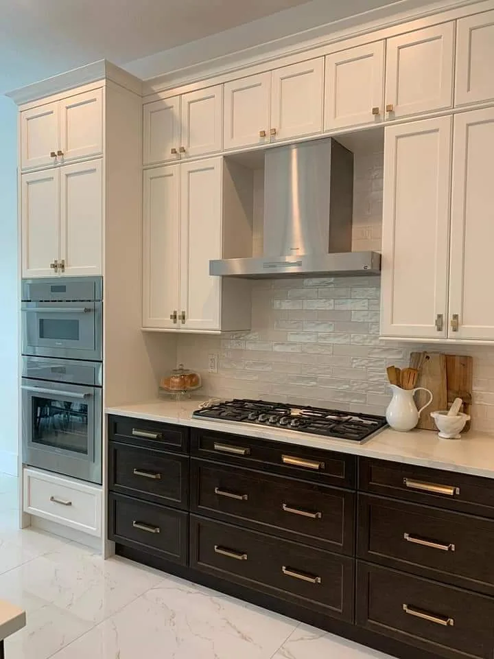 Cocina moderna con gabinetes bicolor: frentes superiores en crema y inferiores en negro, campana extractora de acero inoxidable, estufa de gas, azulejos tipo metro en salpicadero y encimeras de mármol con electrodomésticos integrados.