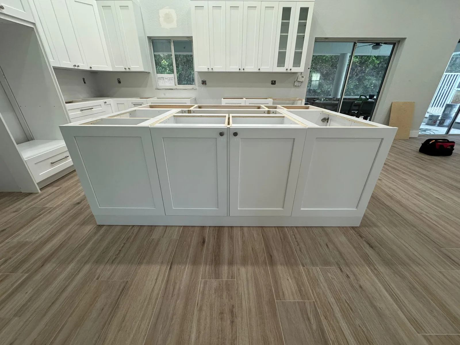 Cocina en construcción con bases de gabinetes blancos instaladas, piso de imitación madera colocado, y gabinetes superiores a lo largo de las paredes con puertas corredizas de vidrio al patio visibles.