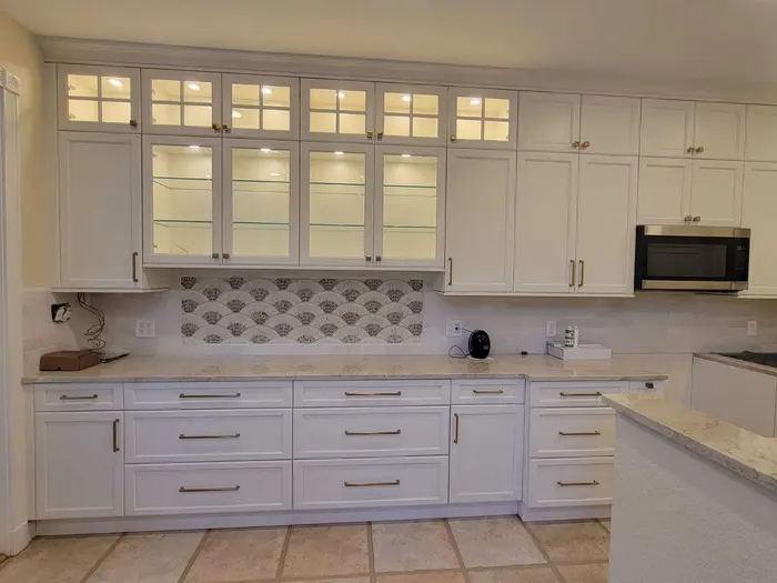 Cocina moderna blanca con gabinetes recién pintados, armarios superiores con frentes de cristal, azulejo de salpicadero con patrón de abanico decorativo y encimeras claras en una casa residencial